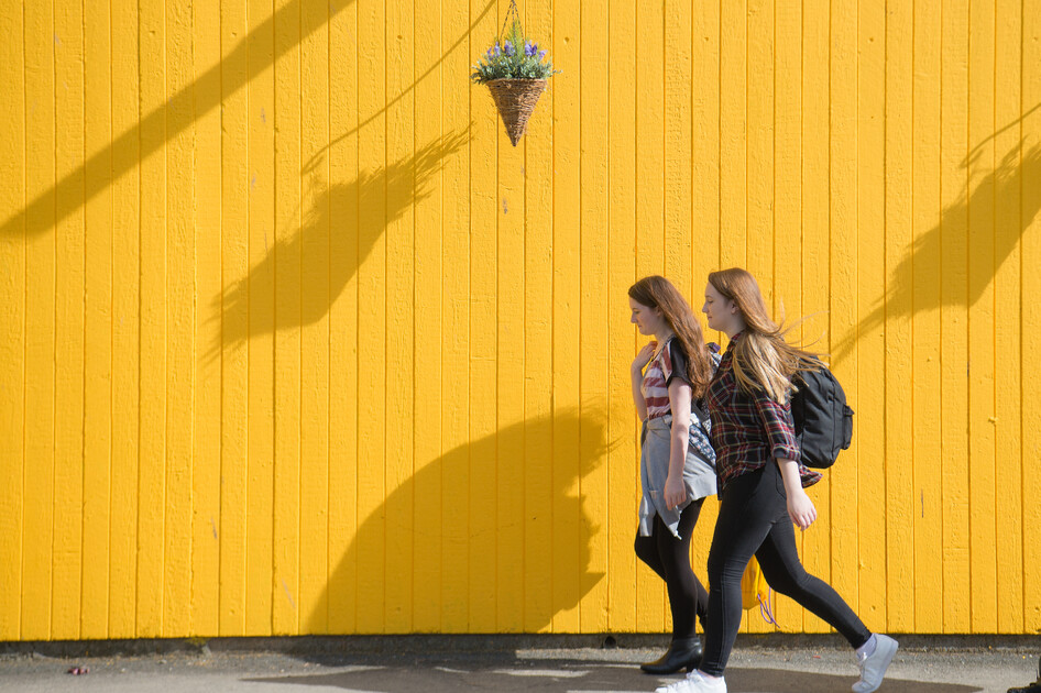 Two students walking together beside a yellow wall