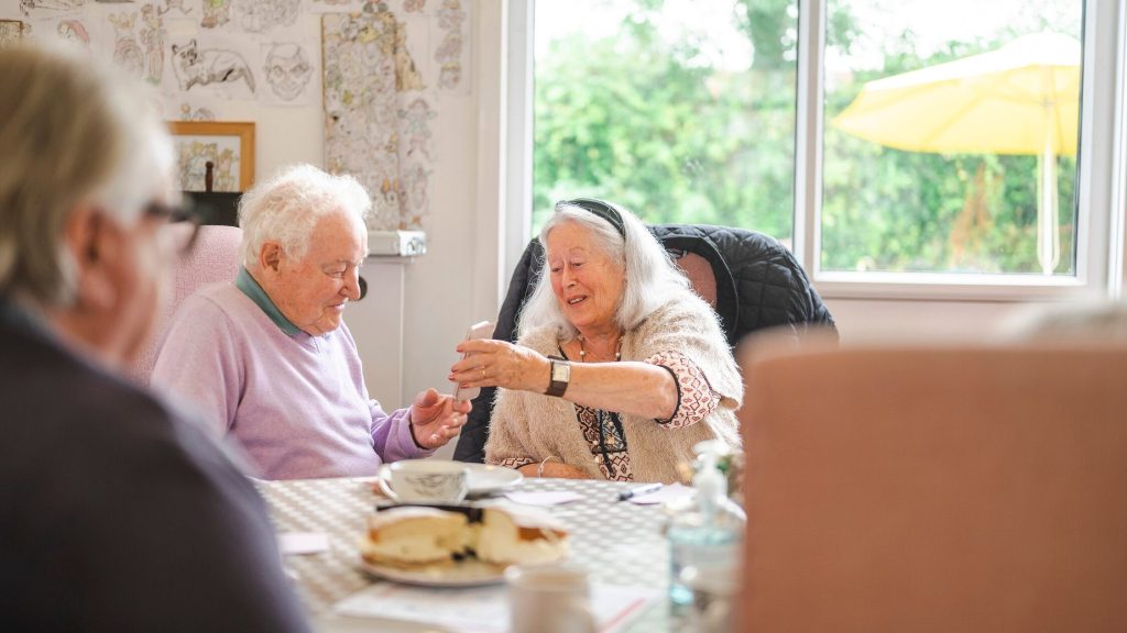 An older woman is showing an older man something on a mobile phone doing a tea morning
